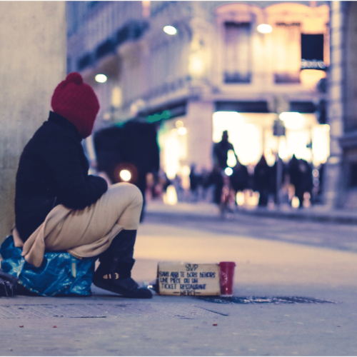 Person without shelter sits with a cardboard sign in a city. Learn how to talk about houselessness with Britt Hawthorne.